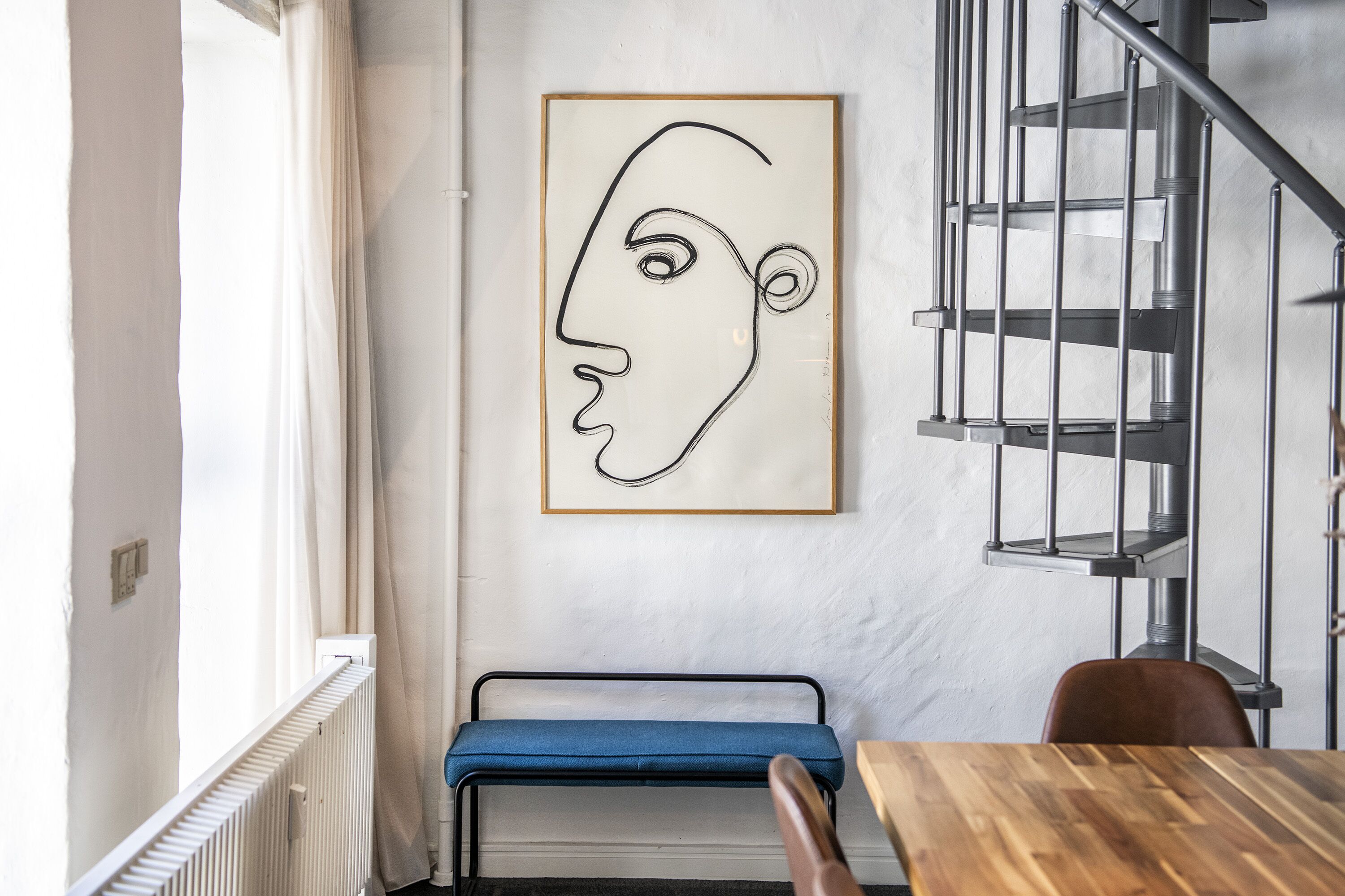 Una habitación con una mesa de madera, un banco azul, una escalera espiral metálica y una obra de arte abstracta enmarcada en la pared.