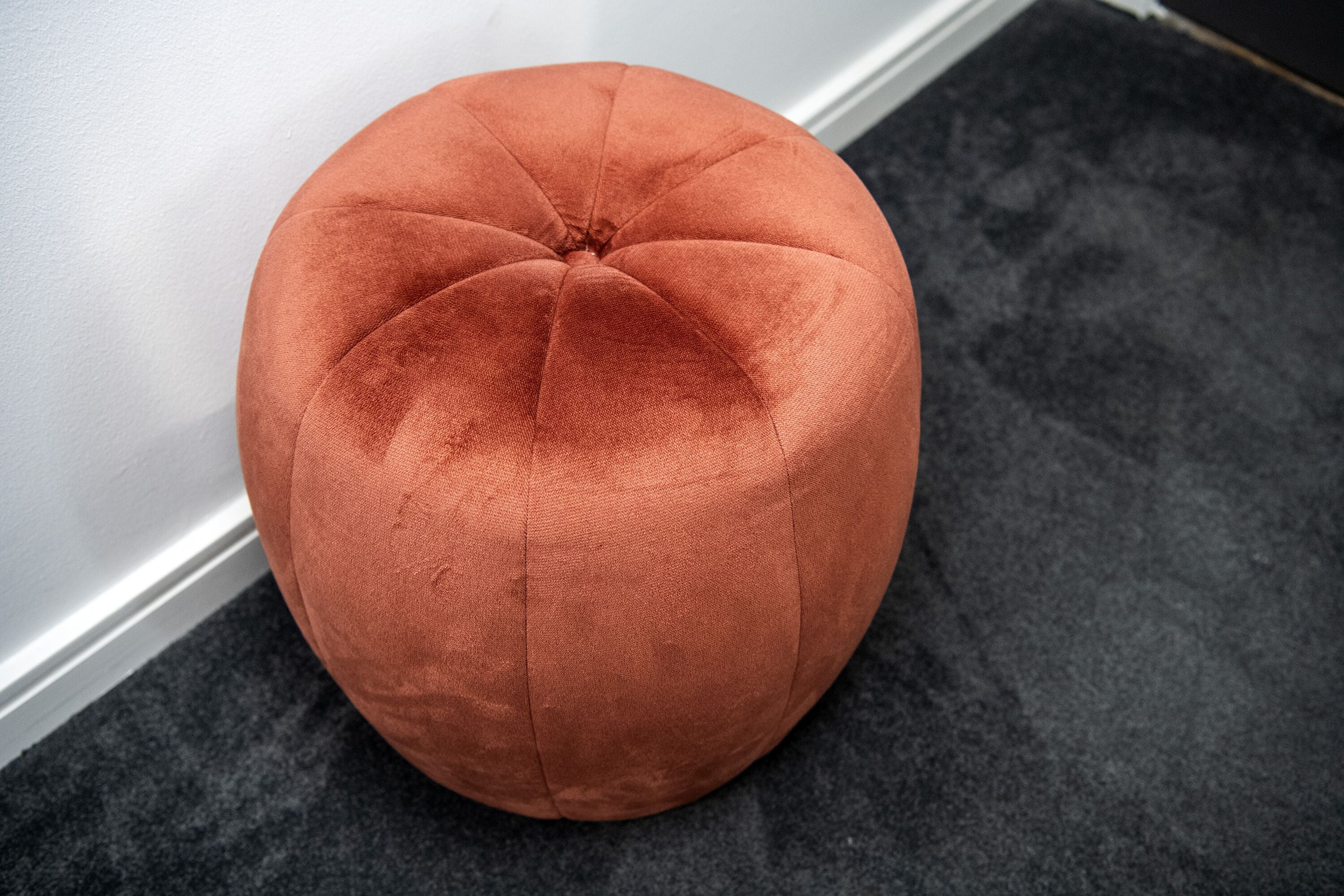 A round, orange ottoman with a tufted design, placed on a dark carpet against a white wall.