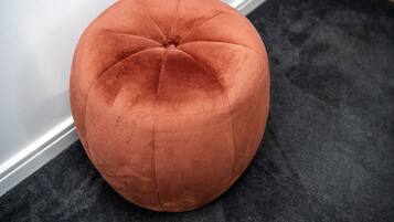 A round, orange ottoman with a tufted design, placed on a dark carpet against a white wall.