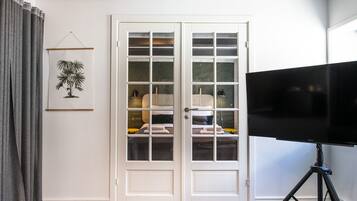 A room with a large flat-screen TV on a tripod stand, a white door with glass panels, and a framed picture of a plant on the wall.