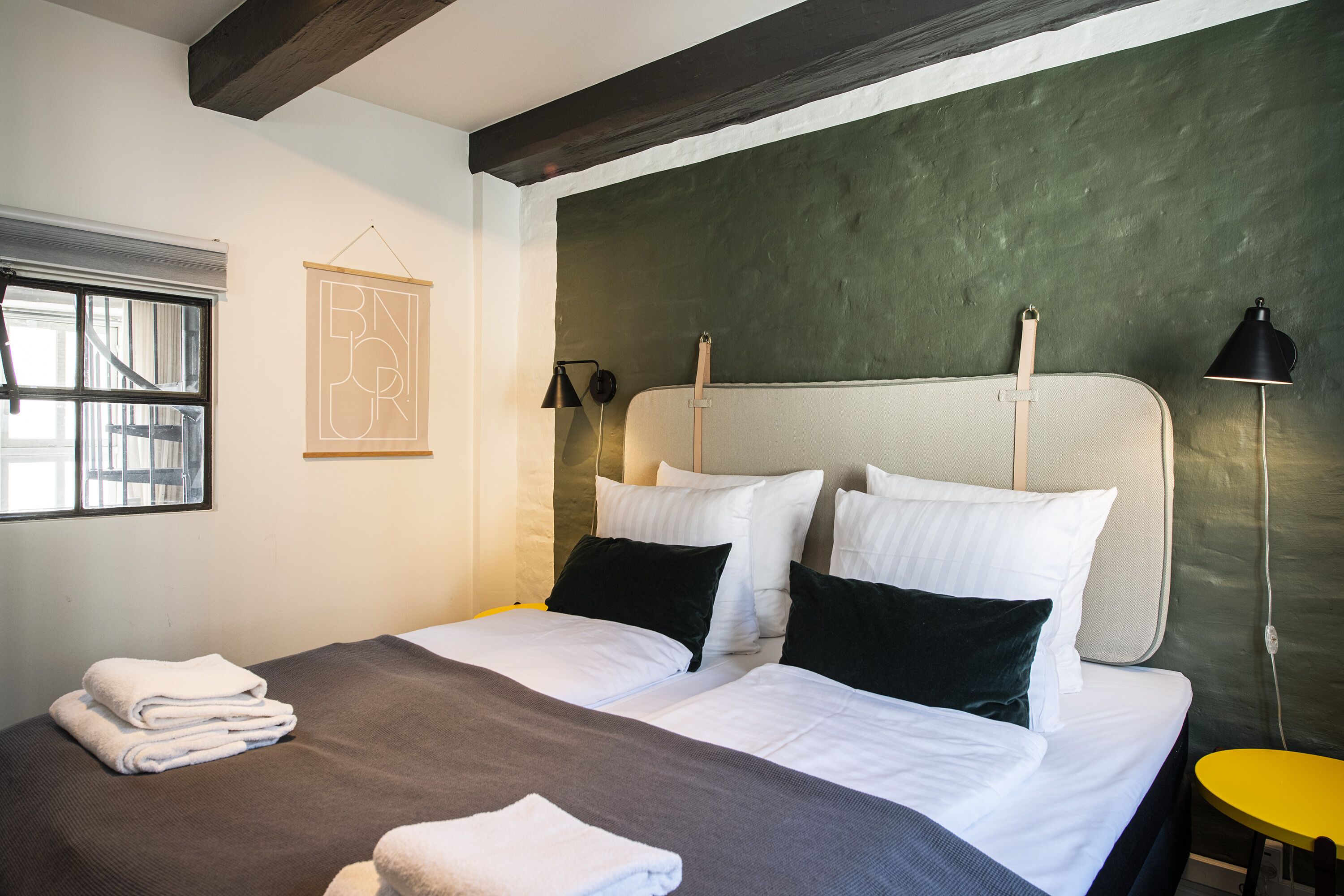 A hotel room with two beds, a window, a green accent wall, and a small yellow table.