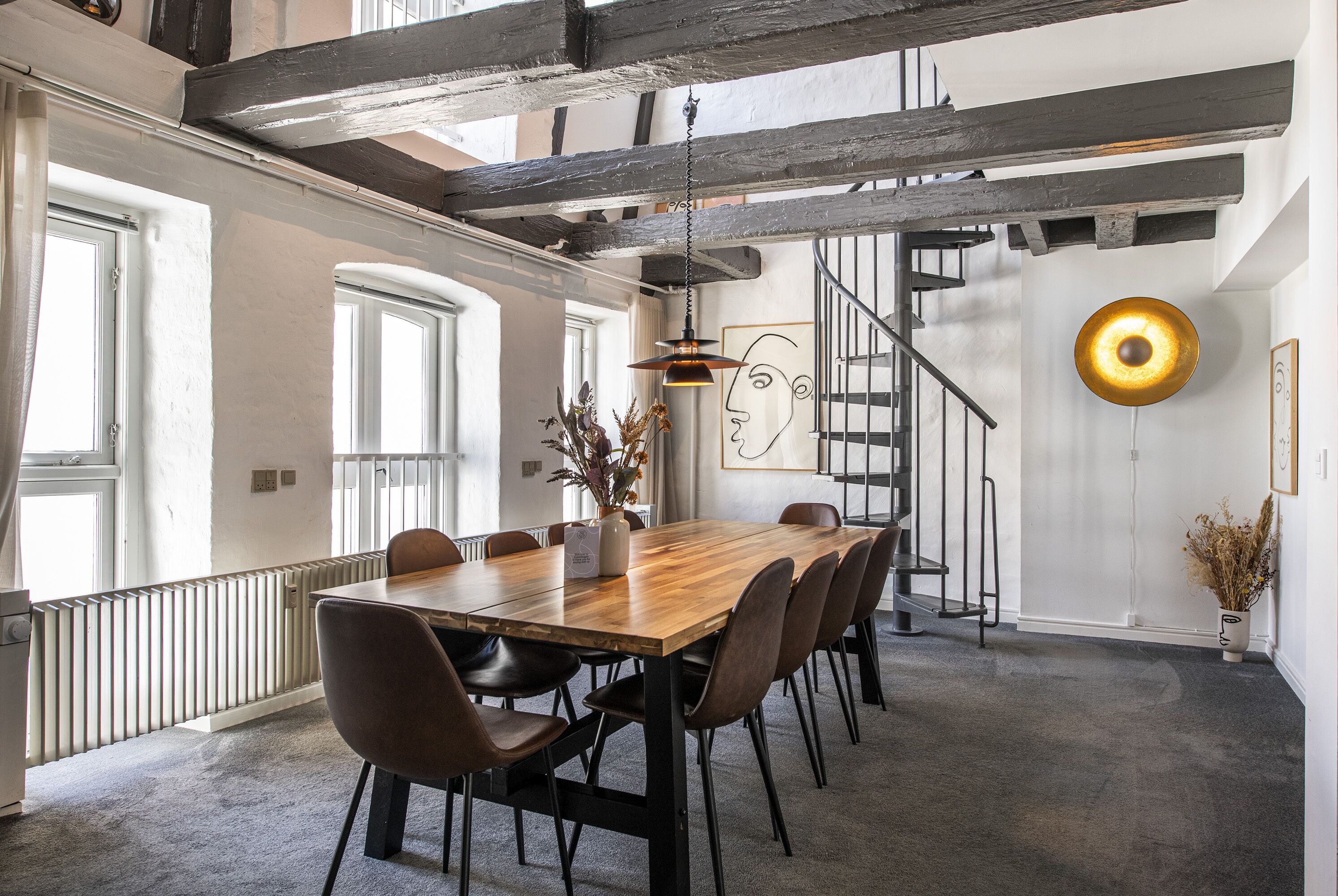 Àmplia zona de menjador amb una taula de fusta llarga i cadires modernes, una escala i una decoració minimalista.