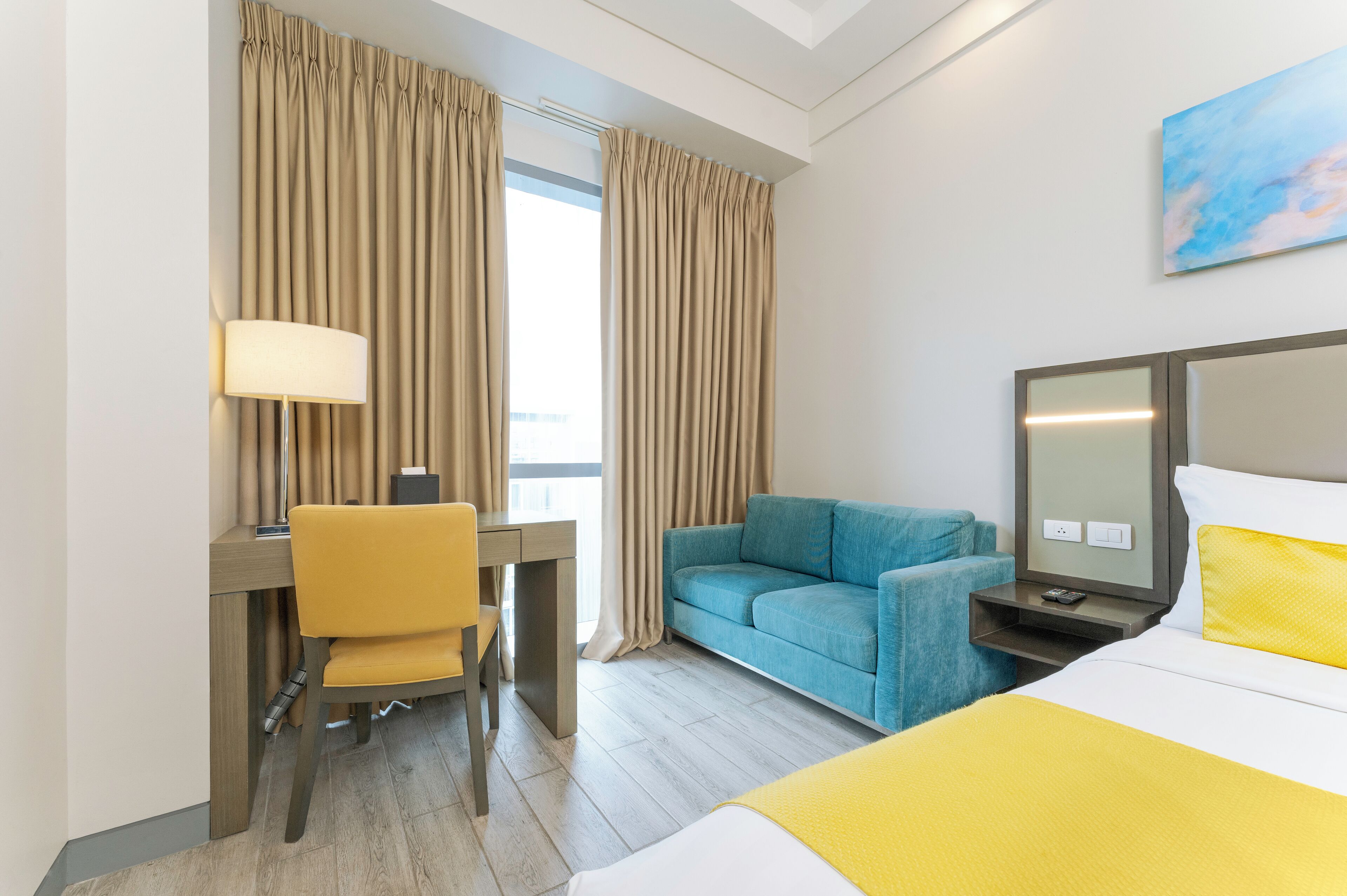 A hotel room with a bed, a desk with a chair, a sofa, a TV, and curtains.