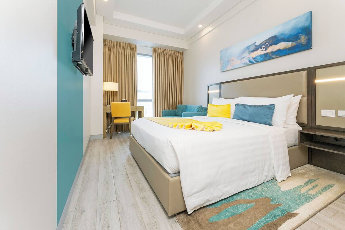 Une chambre d’hôtel avec un lit, un téléviseur fixé au mur, un bureau avec une chaise et une fenêtre avec des rideaux.
