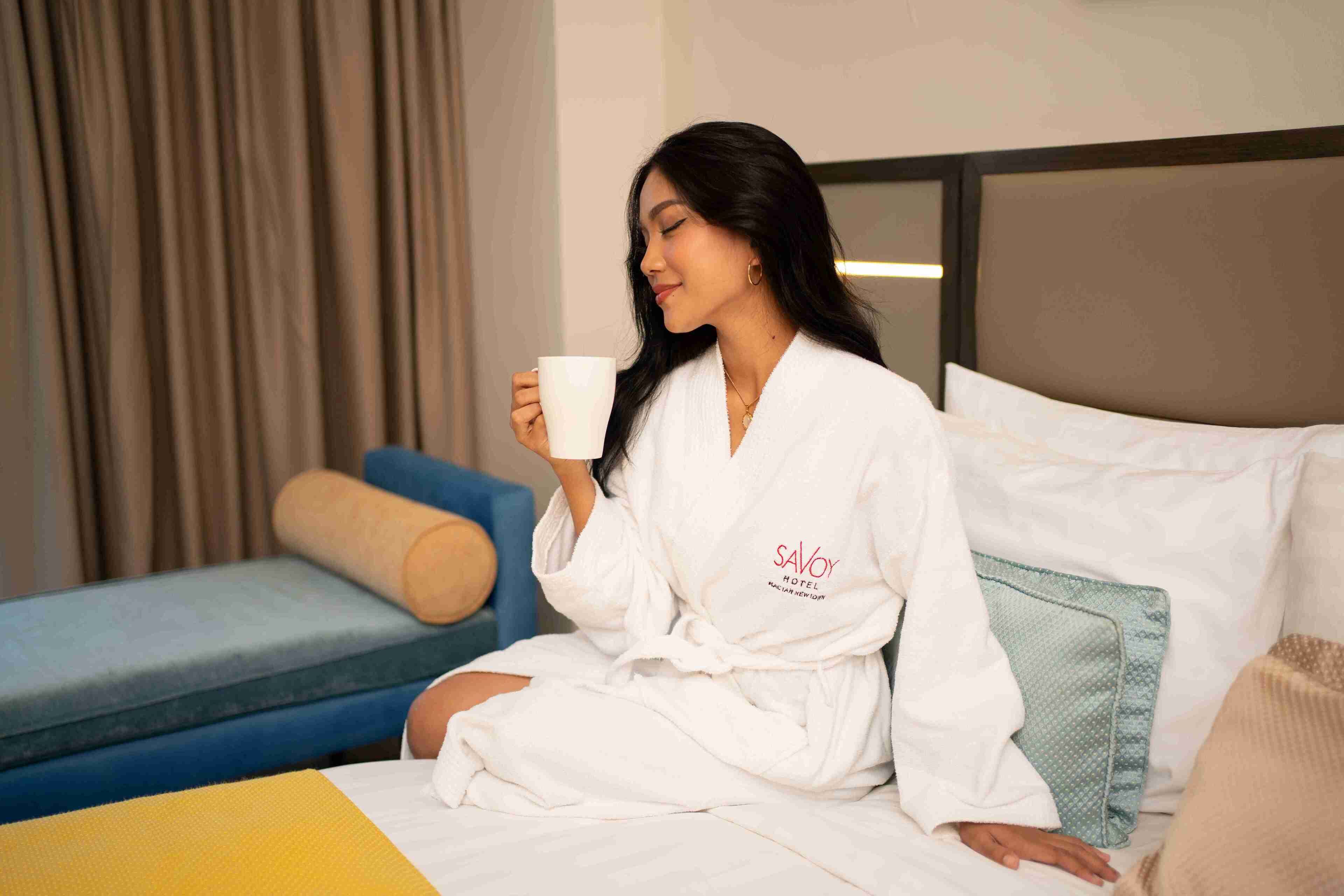 A person in a white robe sitting on a bed with a pillow and a rolled-up towel, holding a white mug.