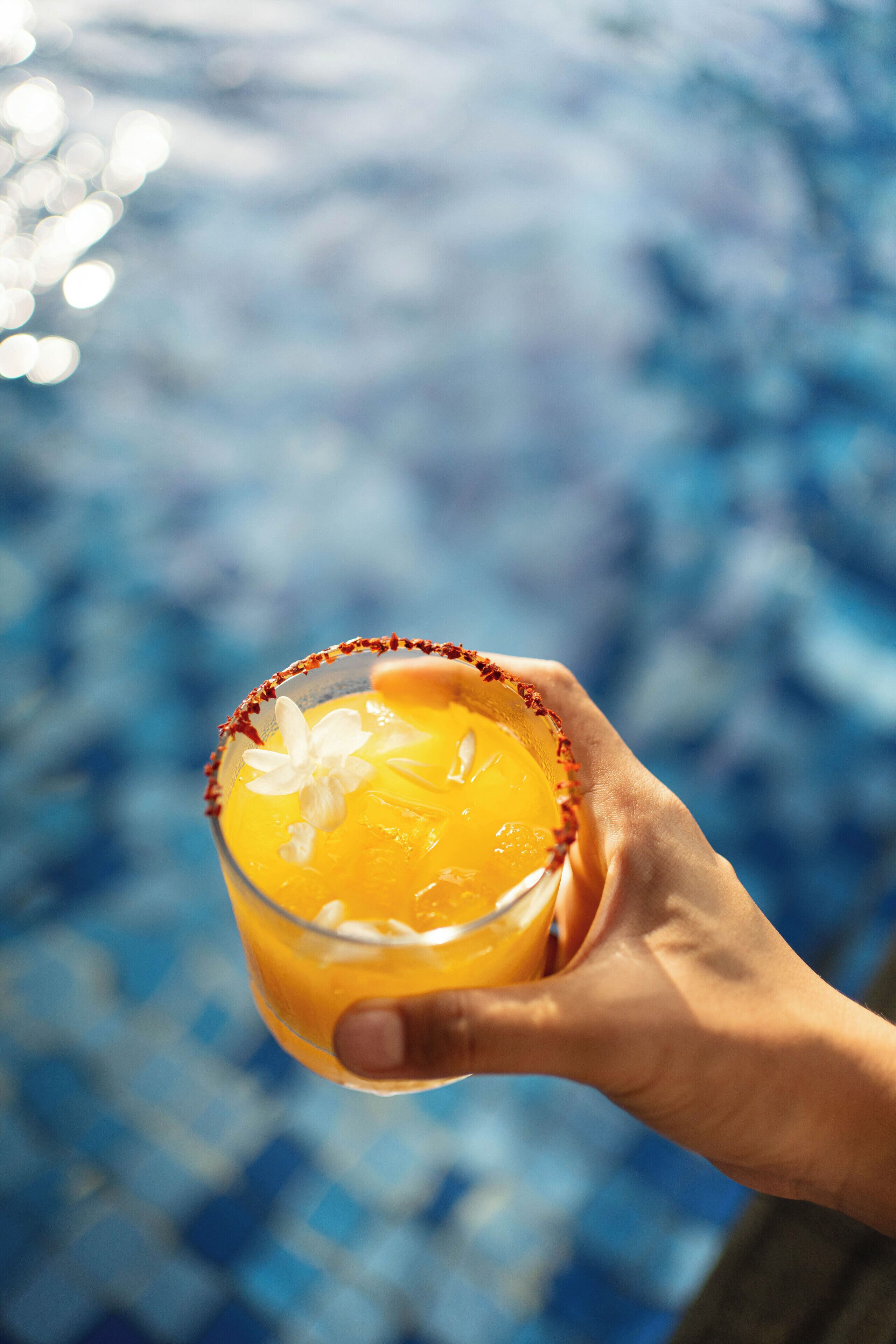 A hand holding a margarita with a salted rim, set against a blurred background of a blue tiled pool.