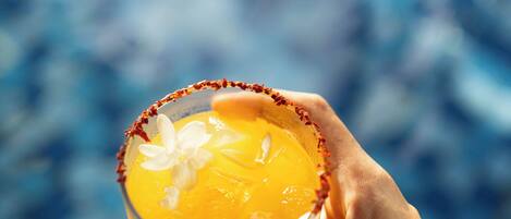 Una mano sostiene una margarita con borde de sal, sobre un fondo desenfocado de una piscina con azulejos azules.