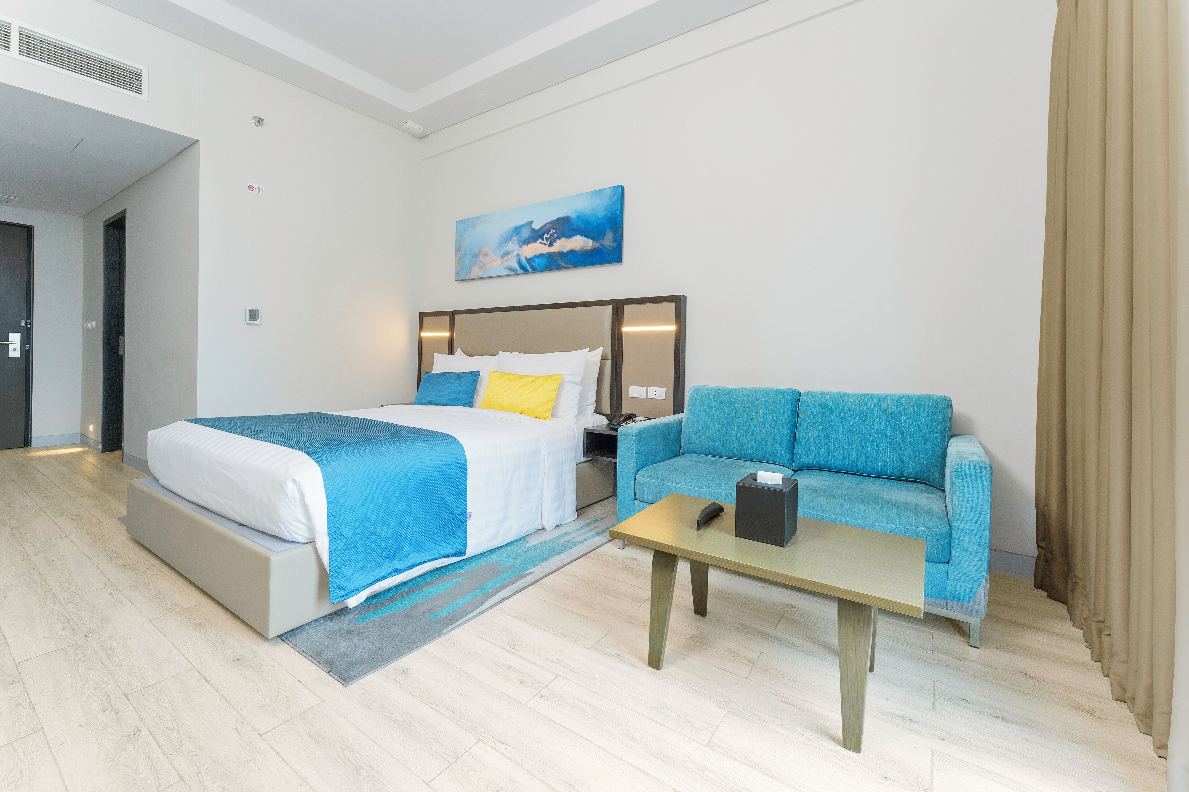 A modern hotel room with a bed, a sofa, a coffee table, and a painting on the wall.