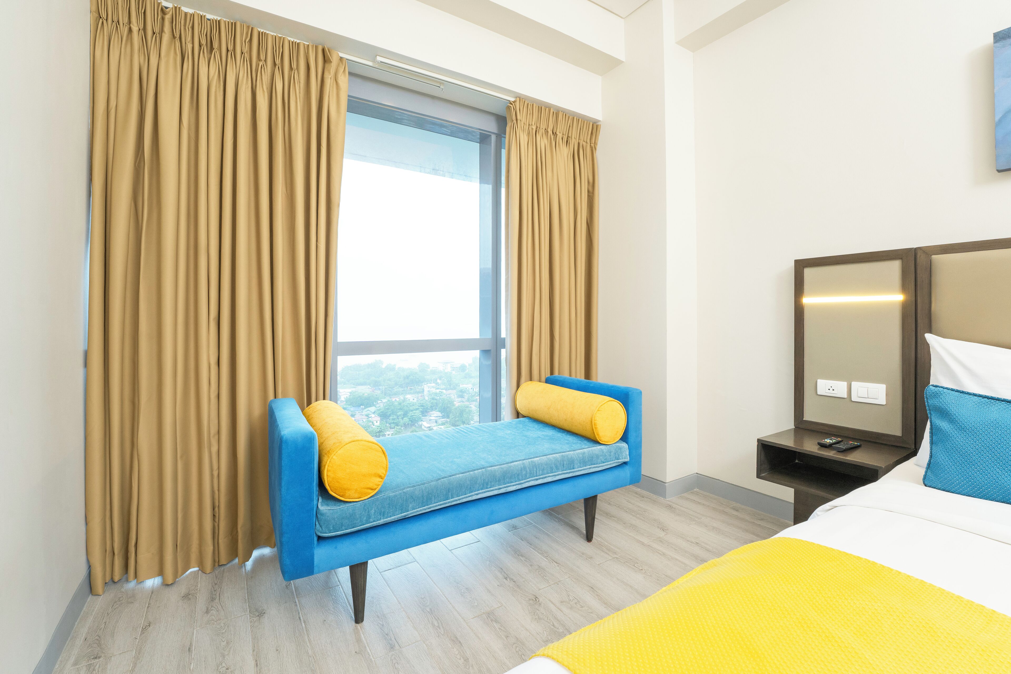 A hotel room with a bed, a blue sofa with yellow cushions, a nightstand, a mirror, and beige curtains.