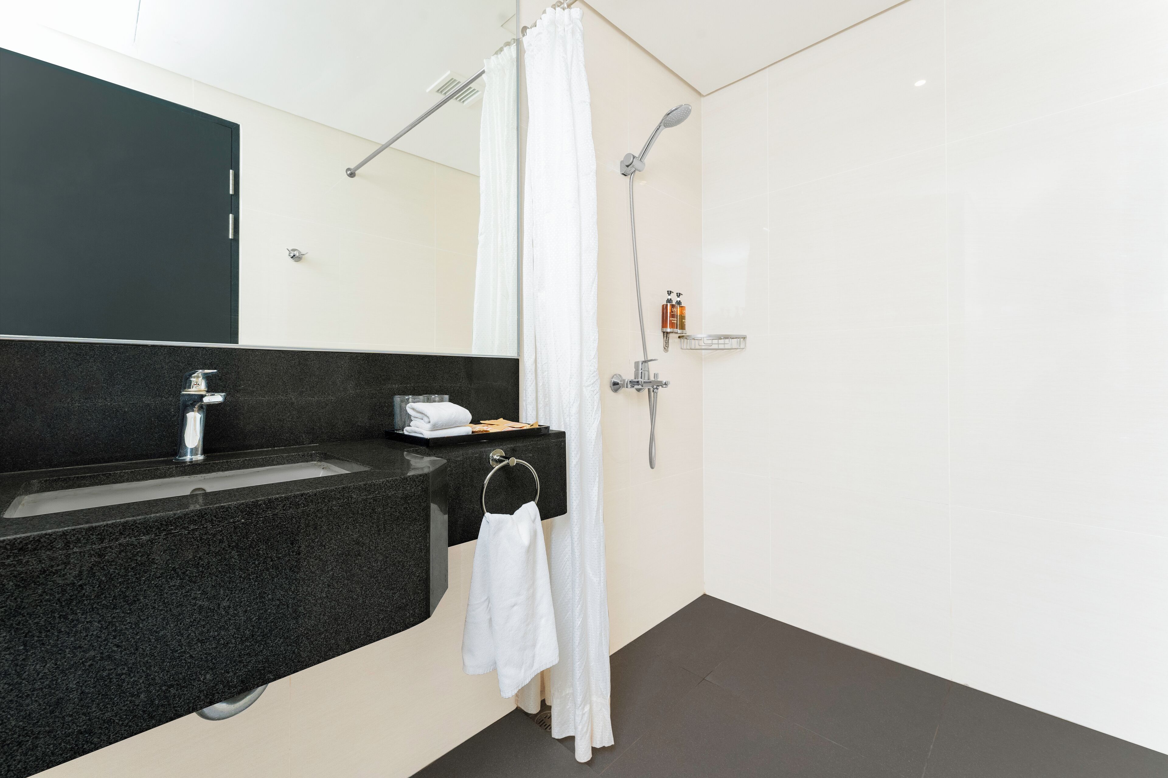 A modern bathroom with a granite countertop sink, a mirror, a shower with a curtain, and a small shelf with toiletries.