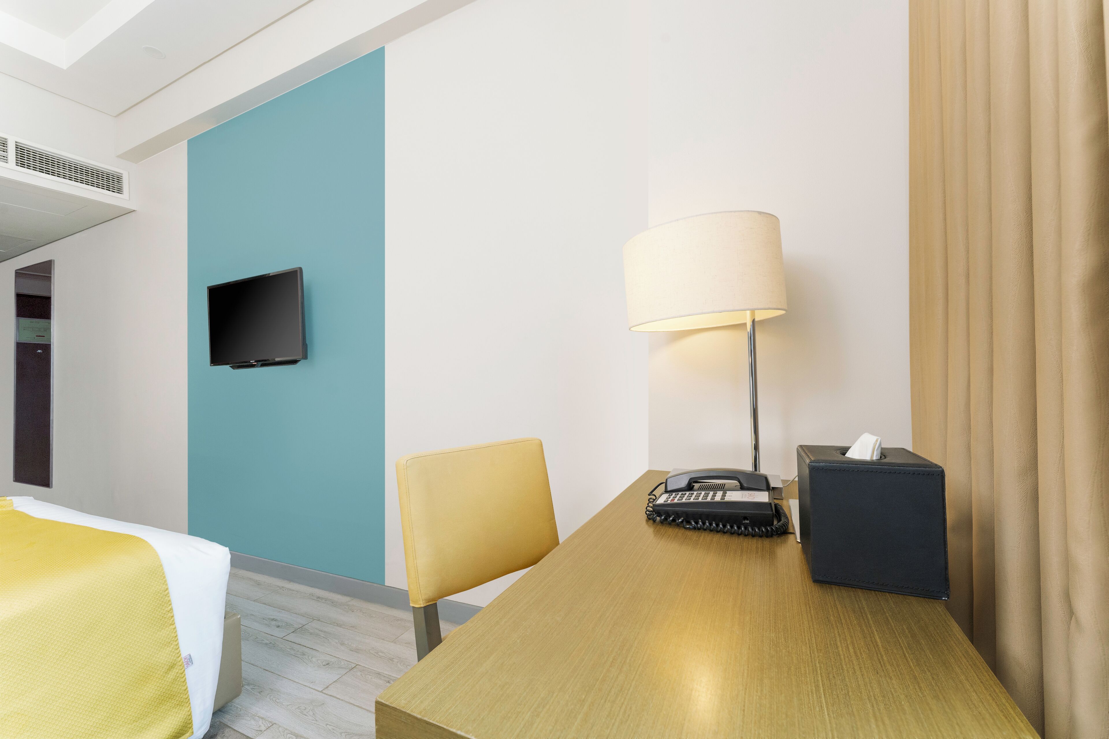 A hotel room with a bed, a desk, a chair, a lamp, a telephone, and a TV on the wall.