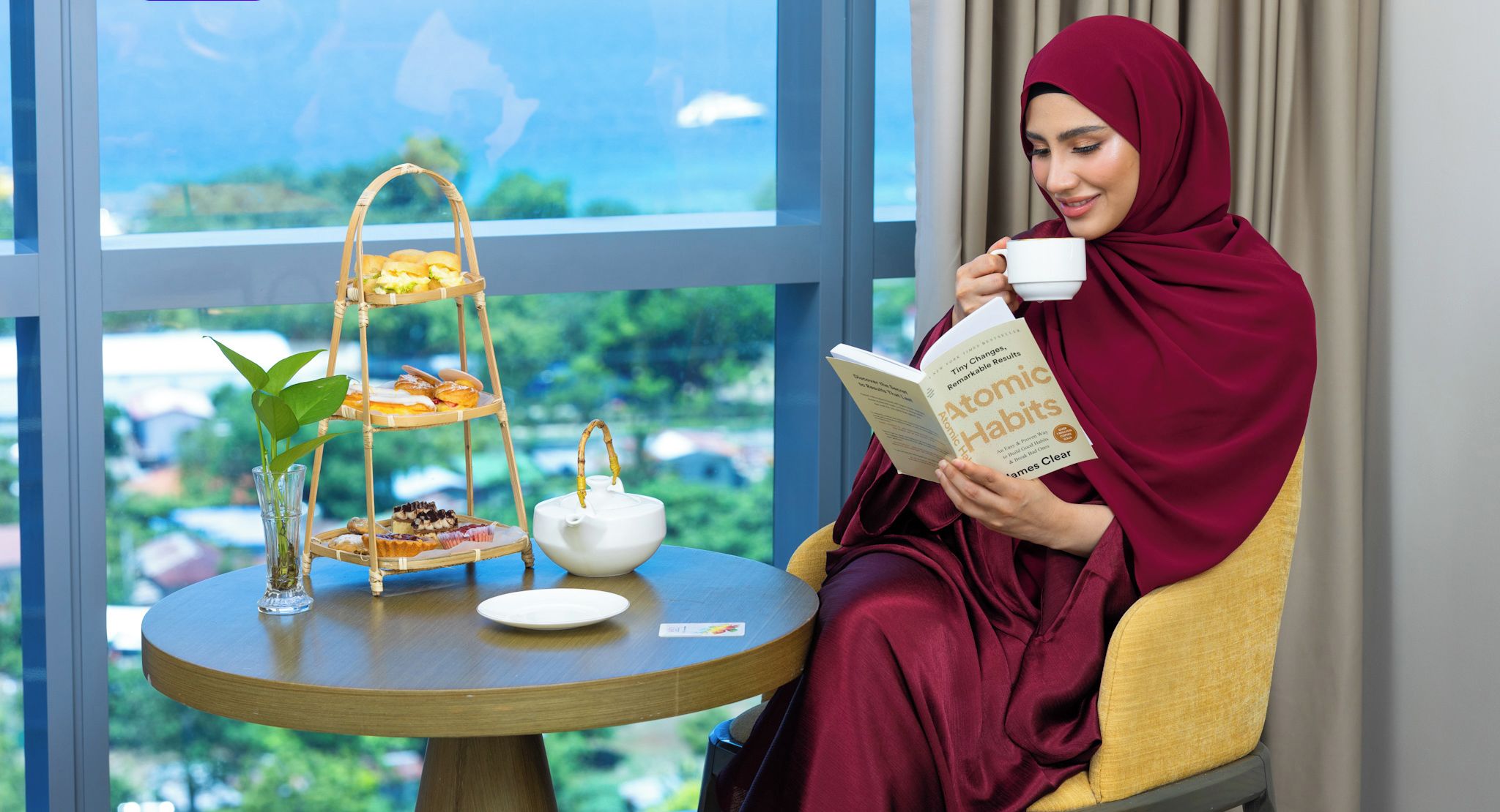A woman reading a book by James Clear in a room with a view of the sea, a table with a tea set, and a tiered tray of pastries.