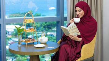 A woman reading a book by James Clear in a room with a view of the sea, a table with a tea set, and a tiered tray of pastries.
