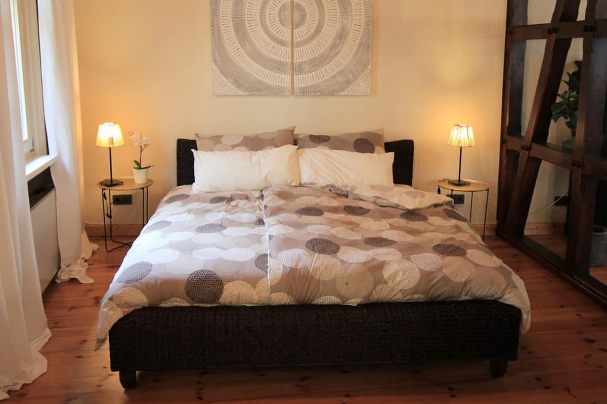 Un lit bien fait, recouvert d’une couvre-lit à motifs, avec deux lampes de chevet et un grand tableau abstrait accroché au mur.