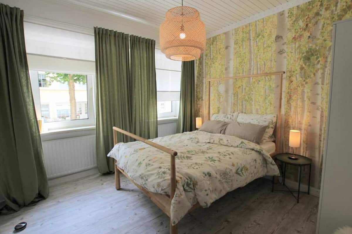 Une chambre à coucher avec un lit en bois, des rideaux verts, un papier peint à motifs et un luminaire suspendu.