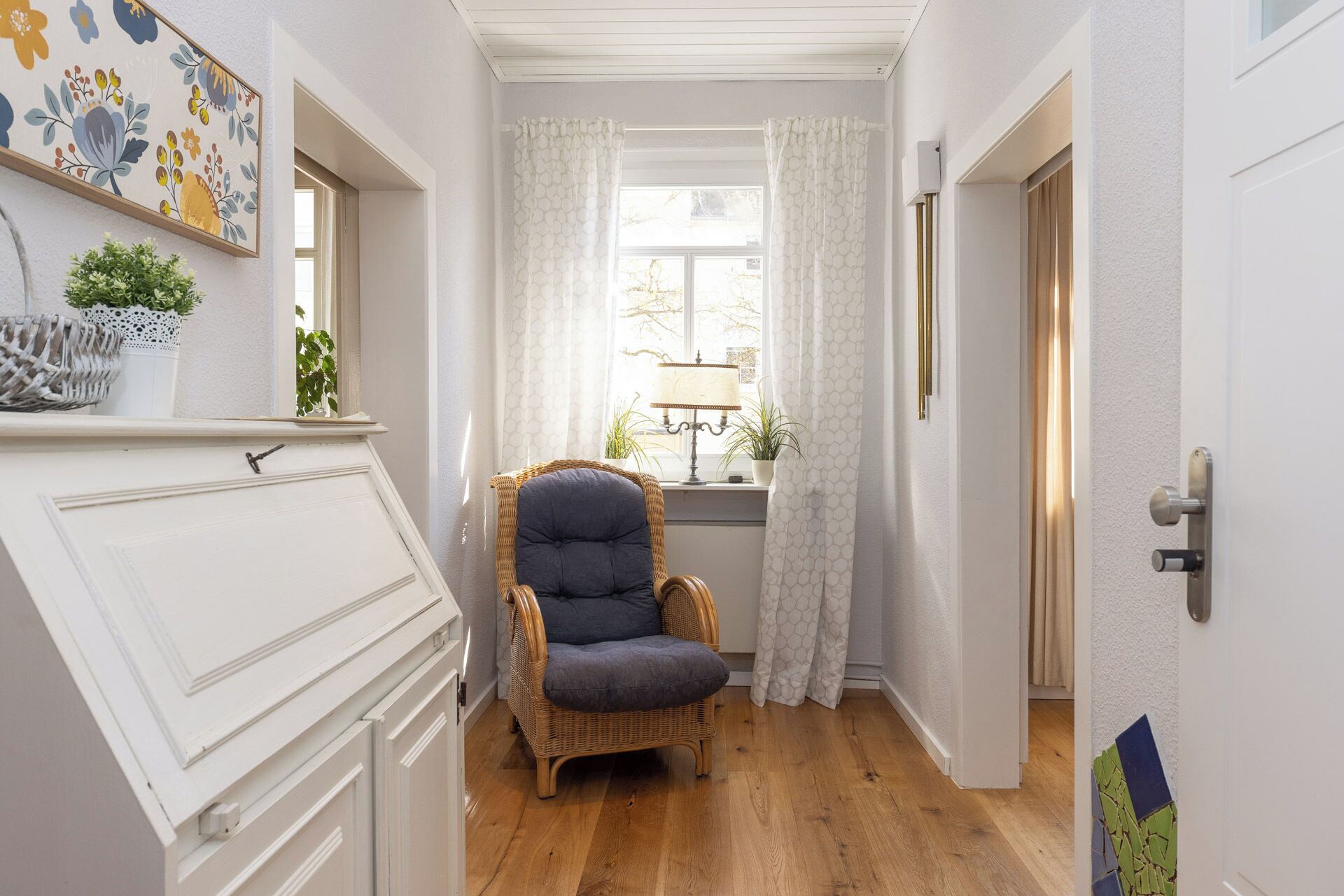 A narrow hallway with a wicker chair, a window with white curtains, and a door with a metal handle.