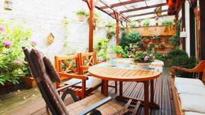 En træterrasse med et rundt bord og stole, omgivet af krukker med planter og en murstensmur.