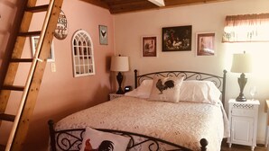 Ein Schlafzimmer mit einer Holzleiter, einem Bett mit einem Kissen in Form eines Hahns sowie Wandbildern, darunter ein Hahn und ein Gemälde von einem Hahn mit Hühnern.