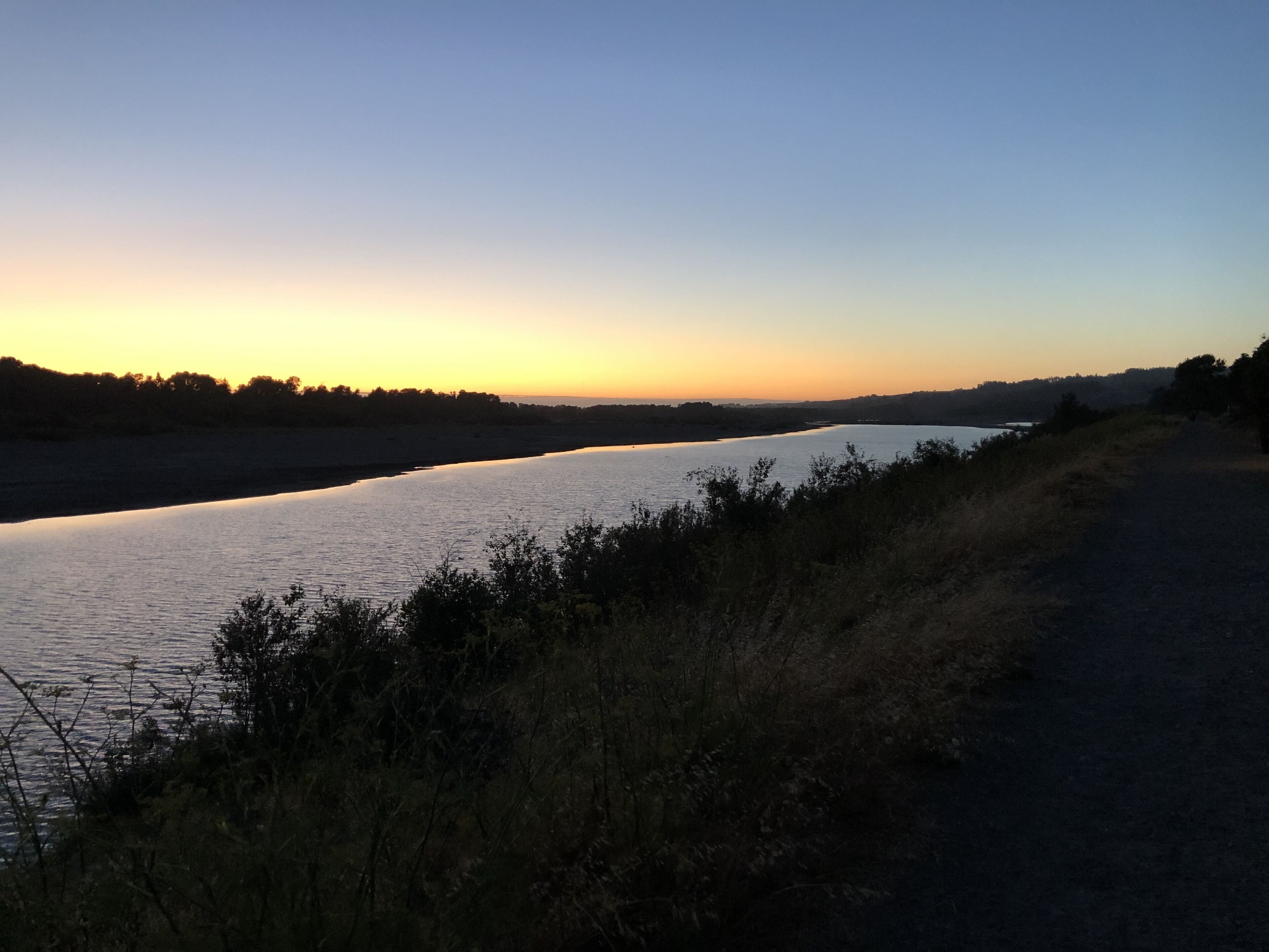 Un río al atardecer con un camino a su lado.