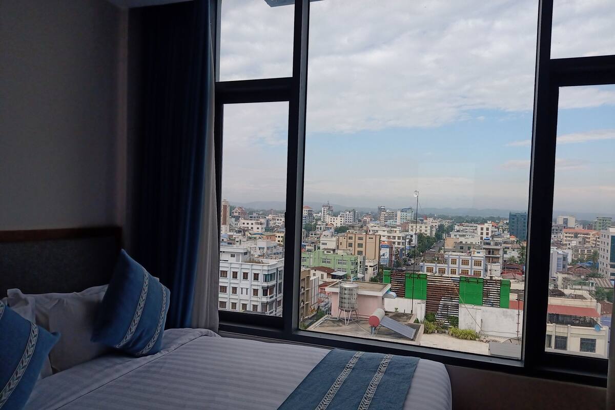 A hotel room with a large bed, a view of a cityscape through two windows, and a city view outside the windows.