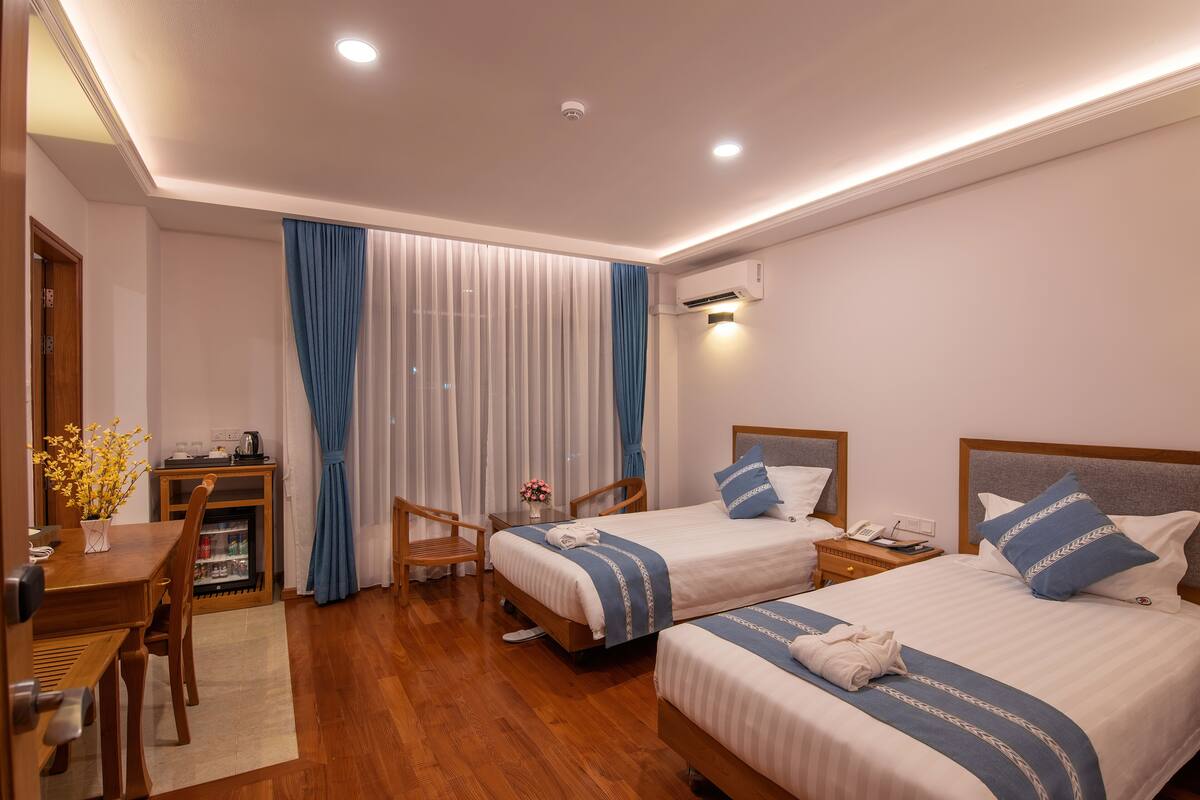 A hotel room with two beds, a desk with a chair, a small table with a vase of flowers, and a minibar.
