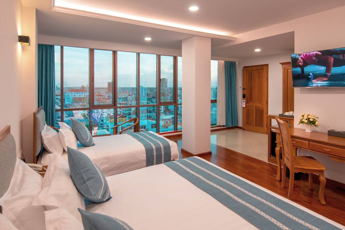 A hotel room with two beds, a desk, a chair, a television, and a large window offering a city view.