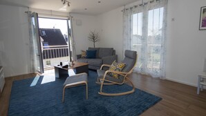 Ein geräumiges Wohnzimmer mit blauem Teppich, einem grauen Sofa, einem Holzschaukelstuhl und einem Couchtisch.
