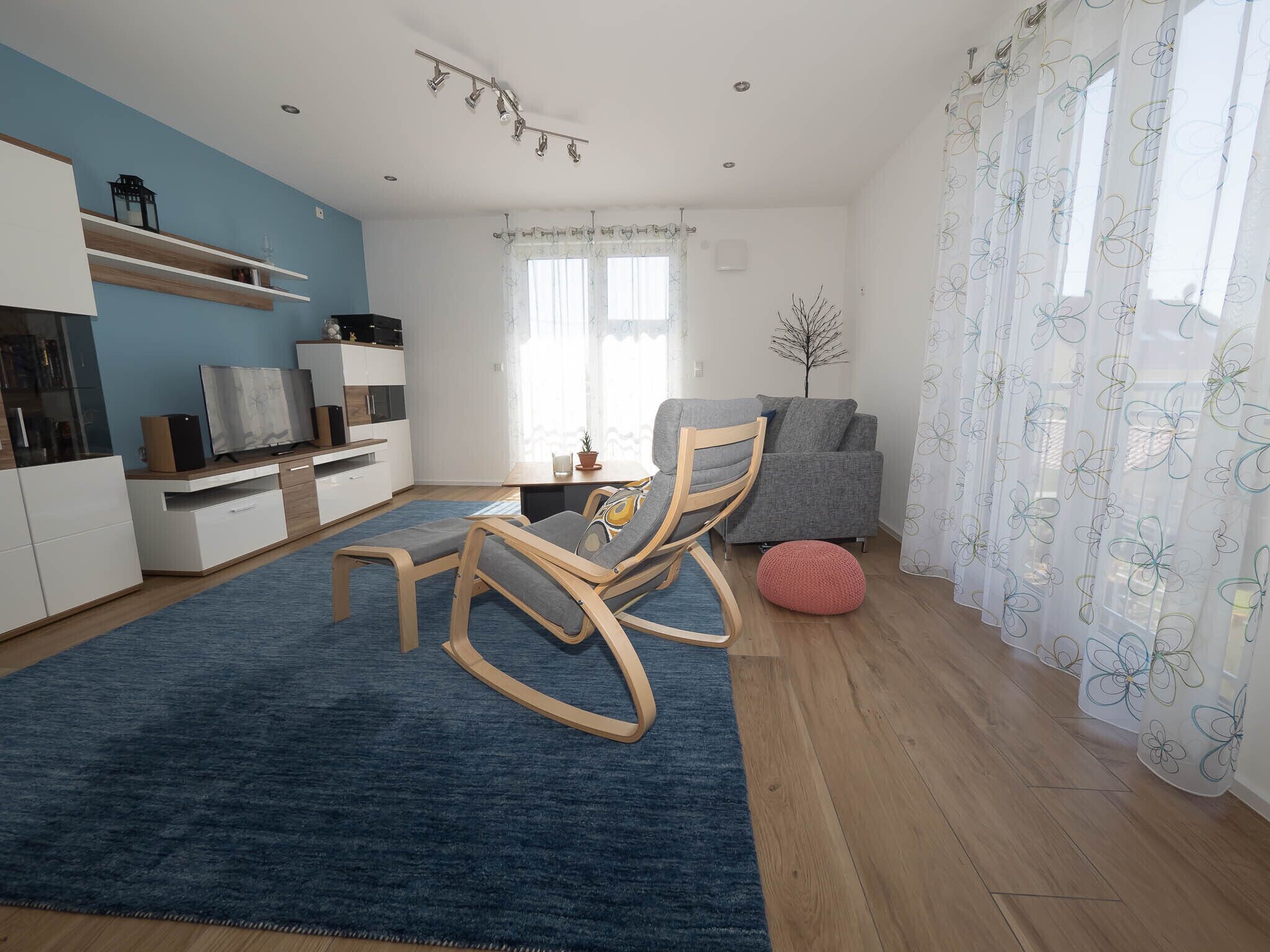 A modern living room with a blue carpet, a wooden rocking chair, a grey sofa, and a red pouf.