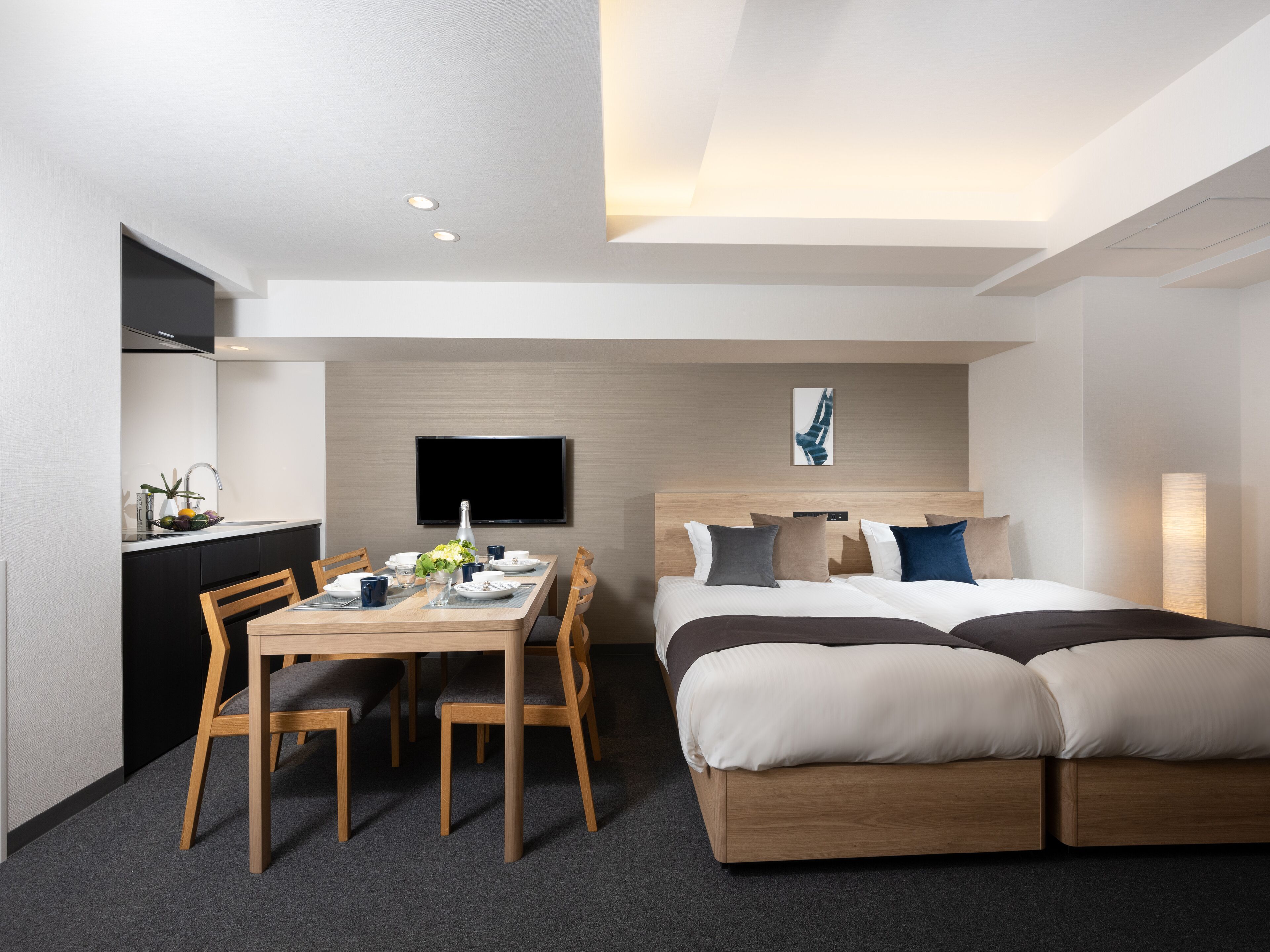 Een moderne hotelkamer met een bed, een eettafel met stoelen, een aan de muur bevestigde televisie en een kitchenette.