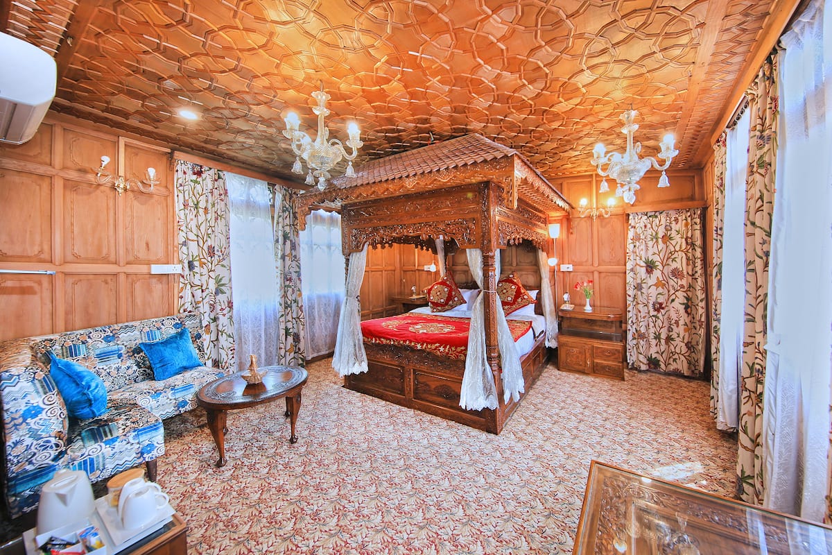Una cama tradicional con dosel y ornamentos de madera, un sofá estampado con motivos florales, y una pequeña mesa redonda con una tetera y tazas.