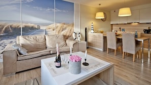 A modern living room with a large window overlooking a beach, a cozy sofa, a dining area with a wooden table, and a kitchen with a clock and pendant lights.