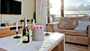 A modern living room with a large window, a flat-screen TV, a sofa, and a table with a bottle of champagne and a candle.