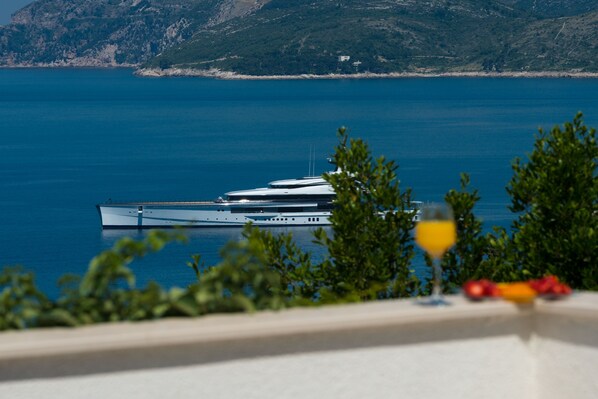 A view of a yacht docked at a marina with a clear blue sea and a mountainous backdrop.