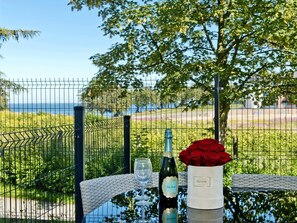 A table with a bottle of wine, a glass, and a vase with red roses, set against a scenic view of a vineyard and the sea.
