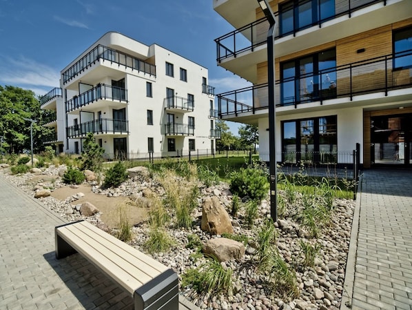 A modern multi-story building with balconies, a landscaped garden with rocks and plants, and a wooden bench.