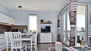 Cozinha moderna com armários brancos, sala de jantar com cadeiras brancas, TV e varanda com cortinas estampadas.