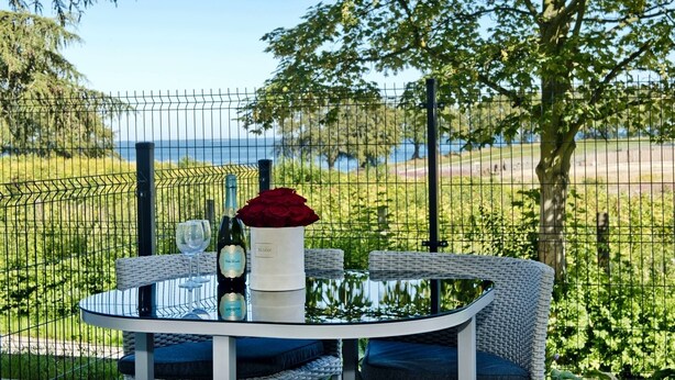 Mesa redonda com uma garrafa de vinho, um copo e um vaso com uma rosa vermelha, com uma paisagem deslumbrante de um lago e árvores ao fundo, delimitada por uma cerca de arame.