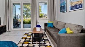 Uma sala de estar moderna com um sofá cinzento, almofadas coloridas, um tapete estampado e vista para uma área externa através de amplas portas de vidro.