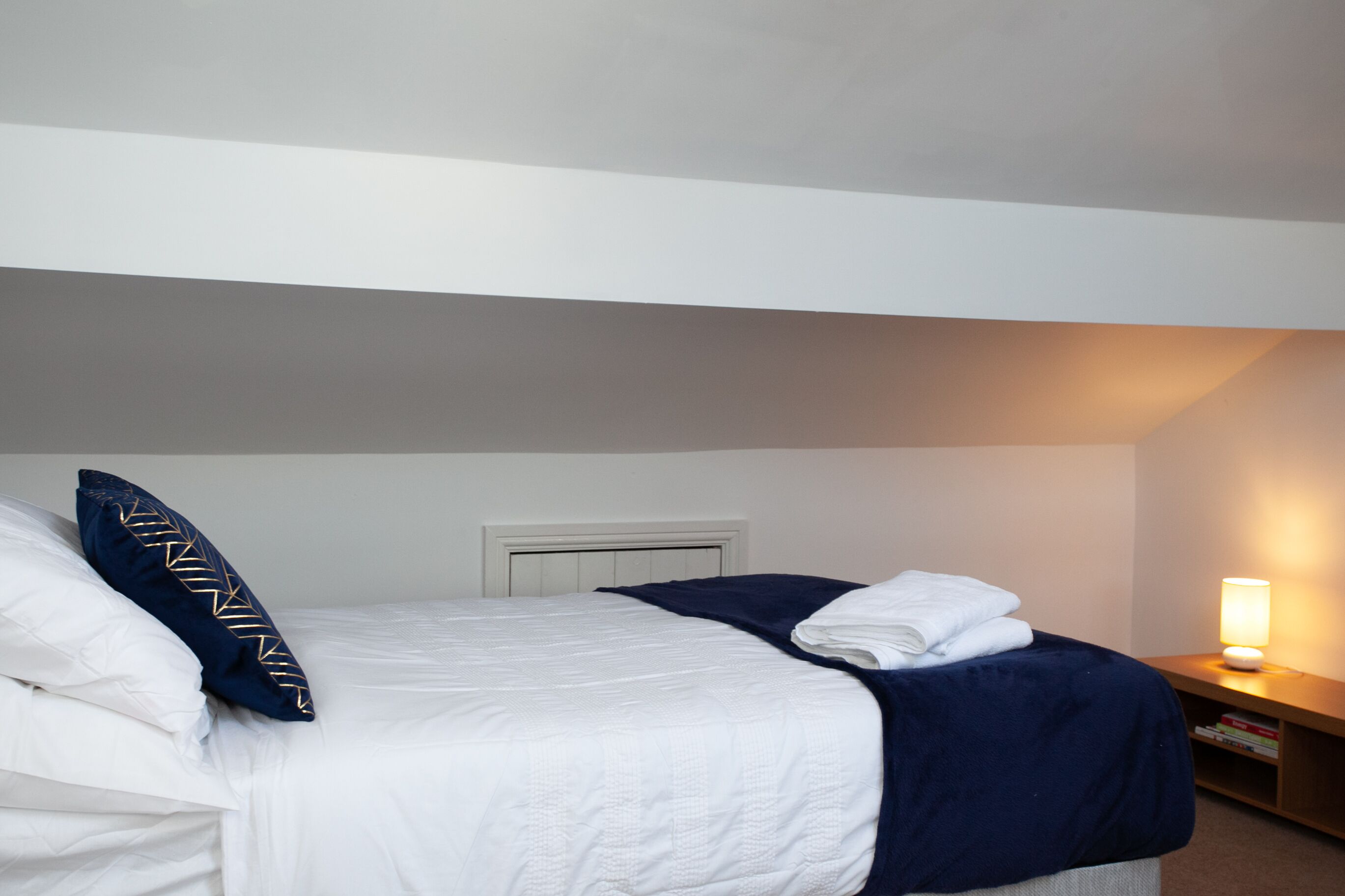A hotel room with a bed, pillows, a bedside table with a lamp, and a wall-mounted heater.