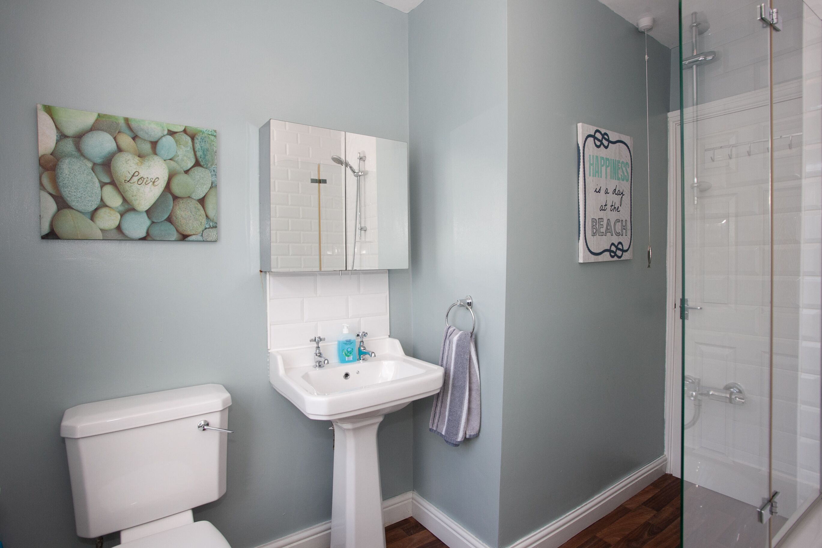 A bathroom with a toilet, sink, and shower. There is a wall-mounted mirror and a decorative wall piece.