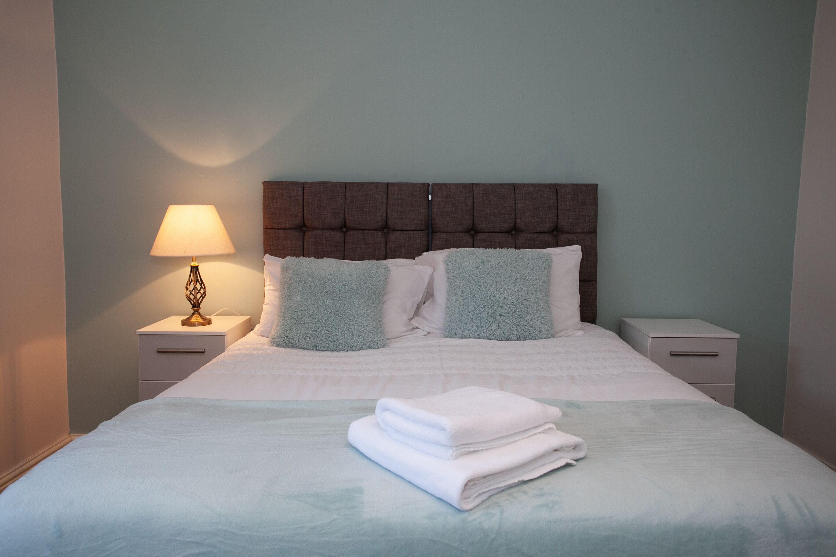 A neatly made bed with a folded towel, a bedside lamp, and a headboard.