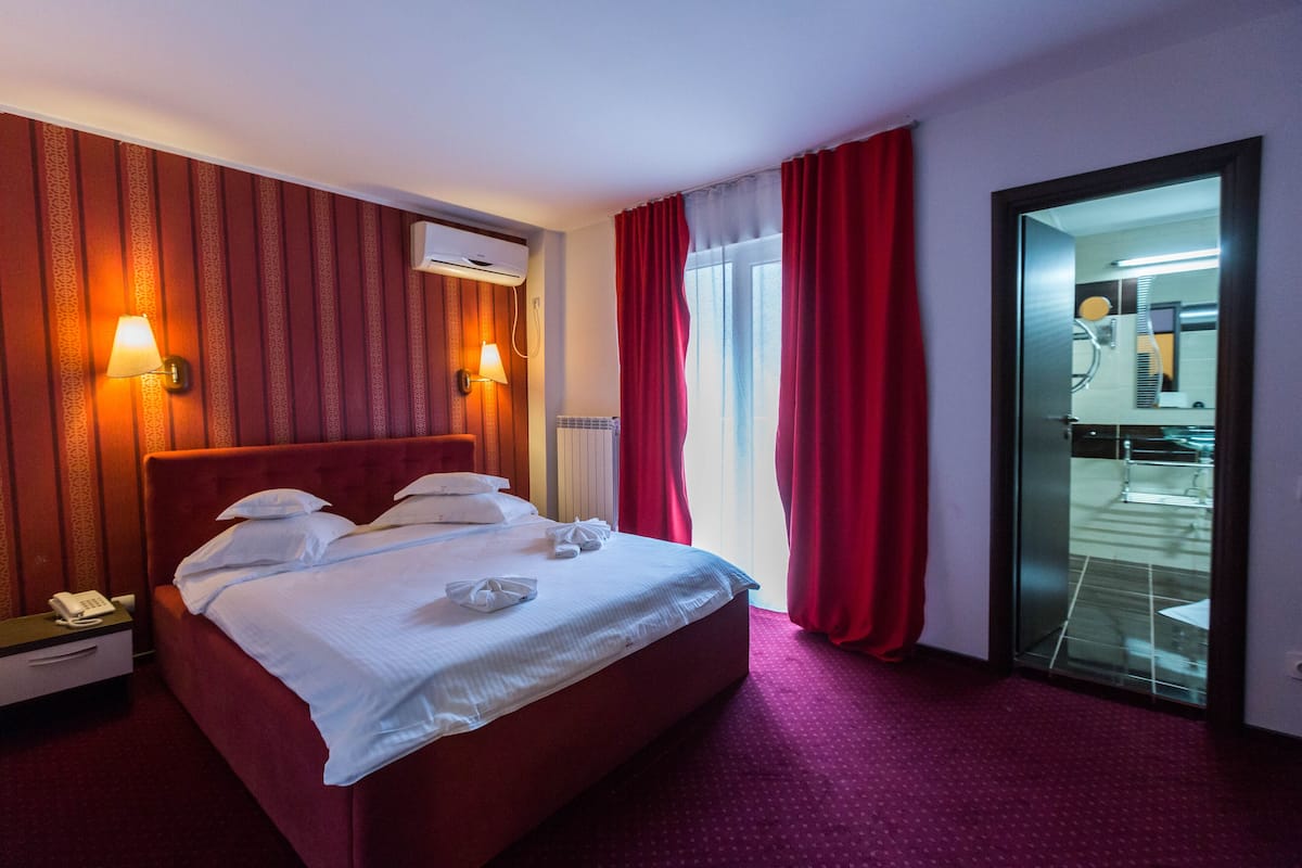 Habitación de hotel con una cama grande, dos lámparas de noche, un teléfono y vistas al baño a través de una puerta de cristal.