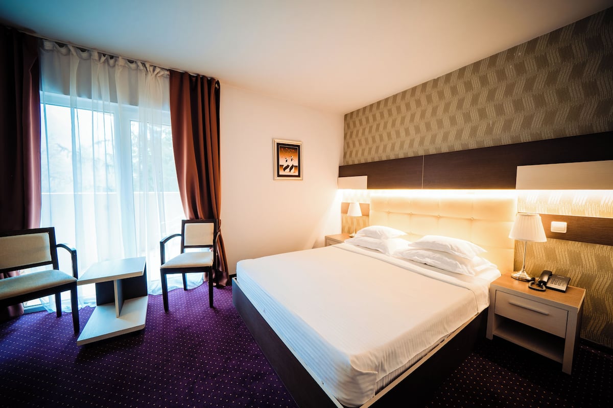 Una habitación de hotel con cama, mesita de noche, lámpara, silla y ventana con cortinas.