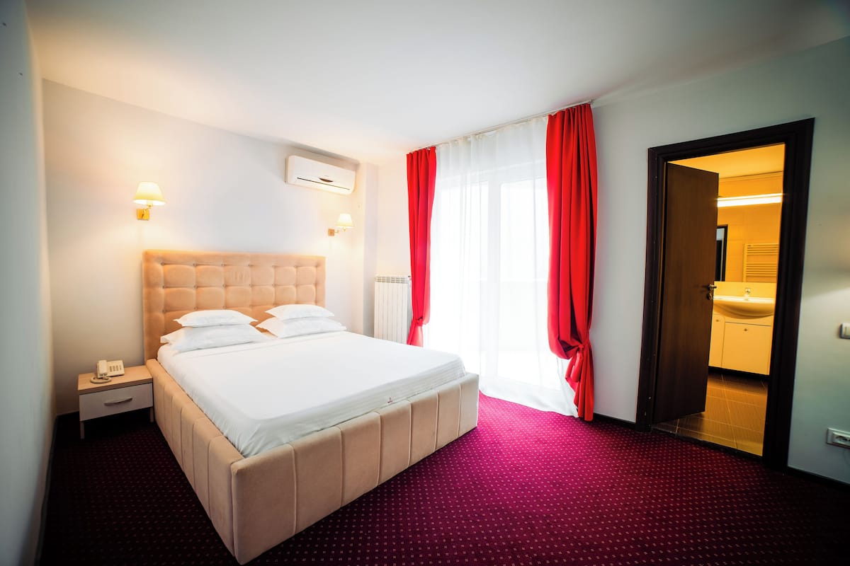 Una habitación de hotel con una cama, una mesita de noche, una lámpara de pared, una ventana con cortinas rojas y una puerta abierta que da a un baño.