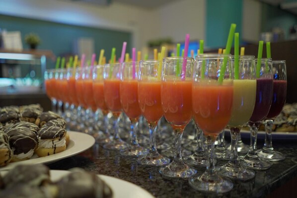 A row of fruit smoothies in wine glasses with colorful straws, and a plate of chocolate-covered pastries in the foreground.