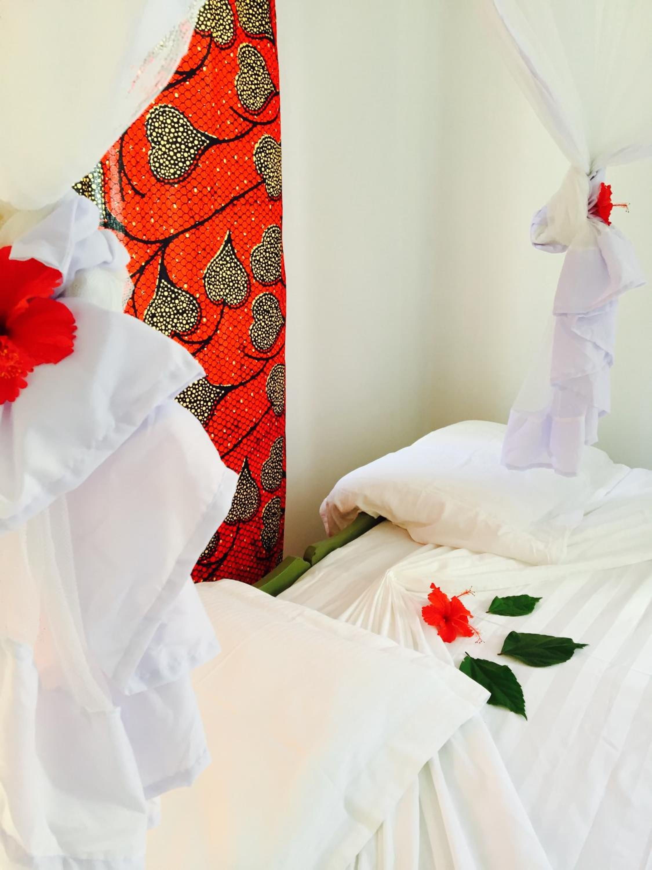 Una cama con ropa blanca y flores rojas, una cortina roja estampada y un cabecero blanco.