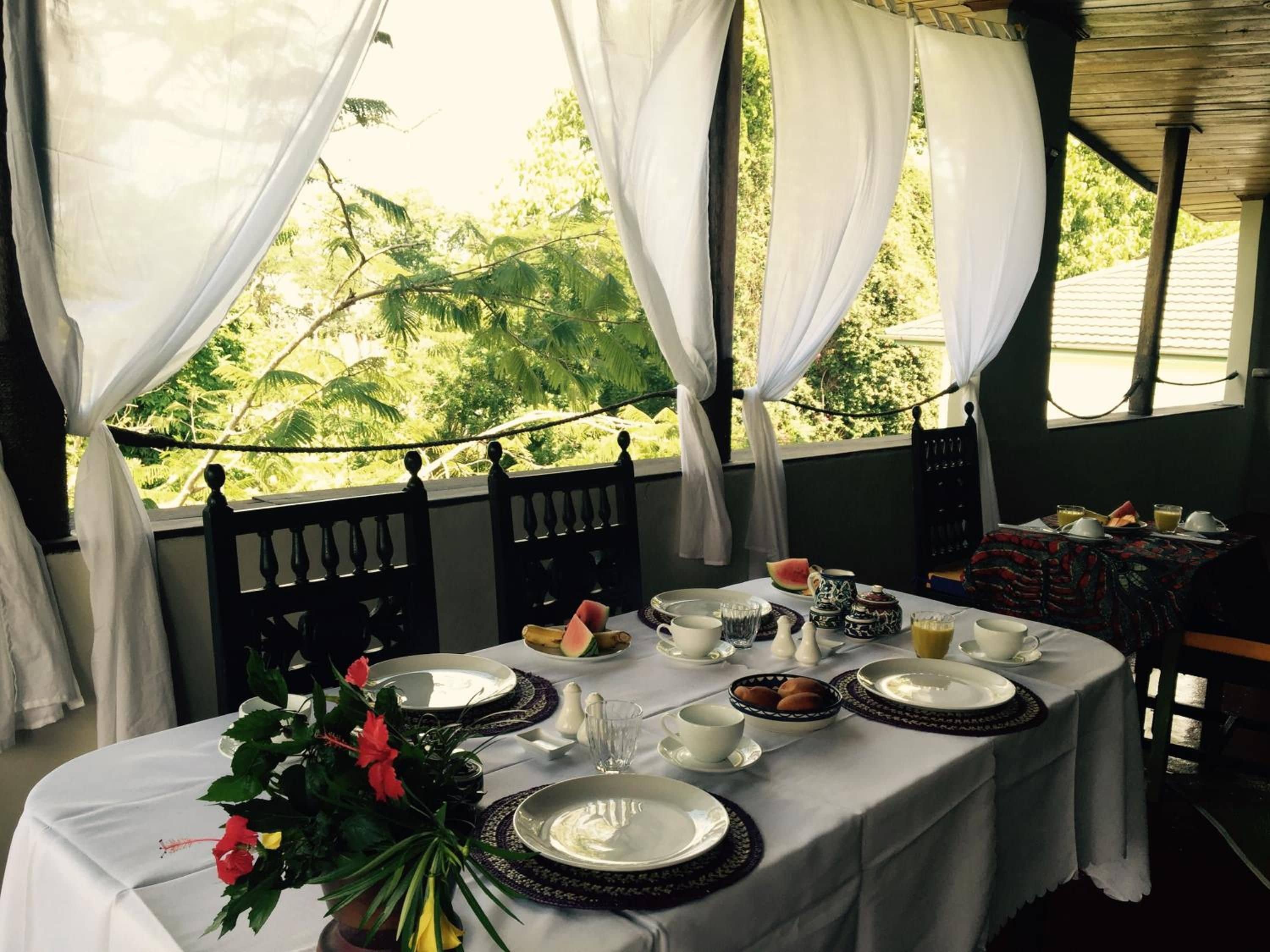A dining table set for breakfast with white tablecloths, plates, and cups, surrounded by a wooden railing and draped white curtains.