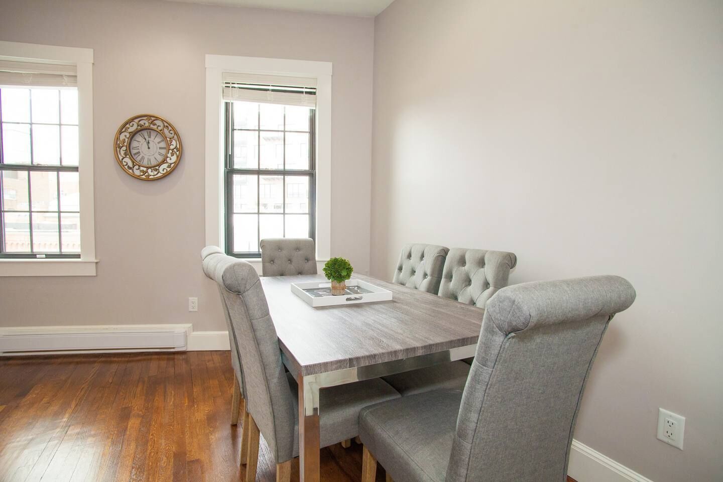 A dining room with a wooden table, grey upholstered chairs, and a decorative clock on the wall.