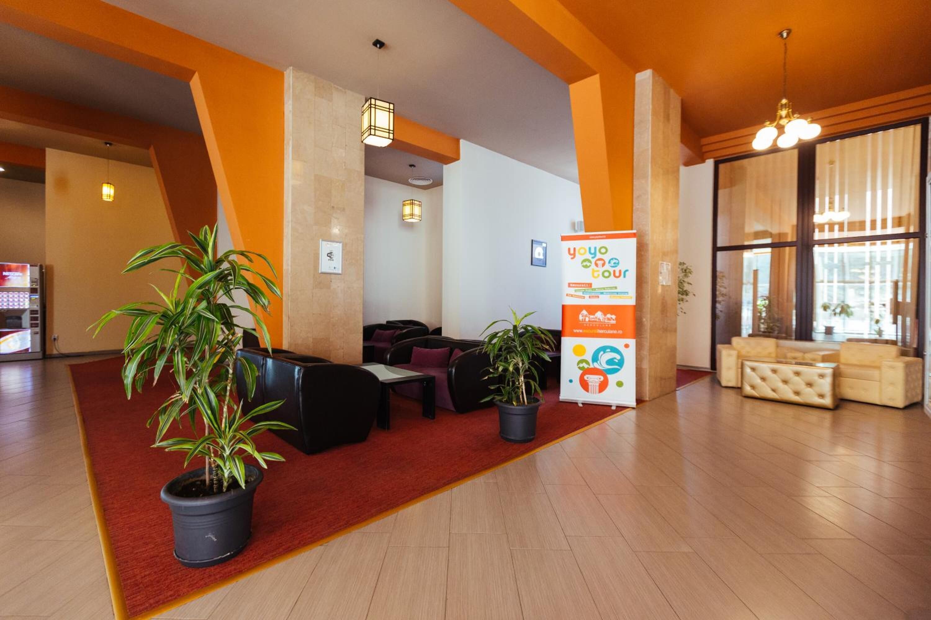 Un hall con alfombra roja, sofás negros, plantas en macetas, un mostrador de recepción y un ventanal con cortinas.