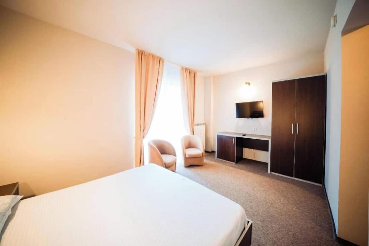 A hotel room with a bed, two chairs, a desk, a TV, and a wardrobe.